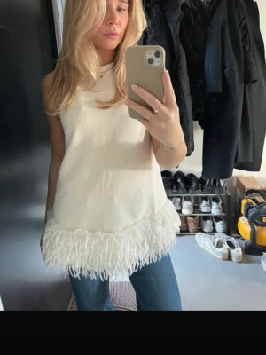 Vit ärmlös topp med fjädrar - Snygg vit ärmlös topp med hög hals och dekorativa fjädrar längs nederkanten. Perfekt för dig som vill sticka ut med en unik detalj. Toppens material känns slätt och lyxigt, och den har en rak passform som ger en stilren look.