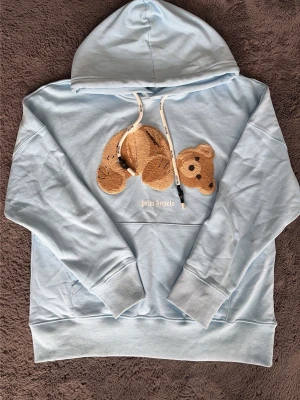 Ljusblå Palm Angels hoodie med björn - Säljer en ljusblå hoodie från Palm Angels med en unik björn-applikation på bröstet och vita dragsnören i huvan. Hoodien har ribbade muddar och nederkant samt en avslappnad passform. Perfekt för dig som gillar streetwear och coola detaljer.