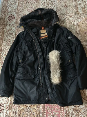 Svart parkas Parajumpers Masterpiece - Säljer en svart parkas från Parajumpers Masterpiece Series Anthology. Jackan har flera stora fickor, huva med avtagbar fuskpäls och detaljer i orange. Perfekt för kalla dagar med sin robusta design och snygga look. Den är äkta och kan bevisas via taggar kvitto finns ej eftersom det var en present 