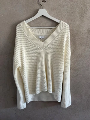 Vit stickad tröja med v-ringning - Snygg vit stickad tröja med djup v-ringning och avslappnad passform. Tröjan har långa ärmar och ribbade detaljer vid halsringning, ärmslut och nederkant. Perfekt att matcha med jeans eller kjol för en enkel och stilren look.