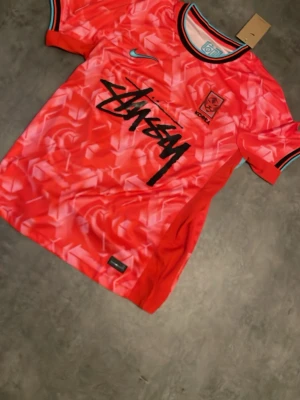 Röd Stüssy x Nike Korea tröja - Snygg röd sporttröja från Nike i samarbete med Stüssy och Korea. Tröjan har ett grafiskt mönster, svart Stüssy-logga på bröstet, Korea-märke och turkosa detaljer. Rund halsringning med svart kant och randiga muddar. Perfekt för dig som gillar streetwear och sport.