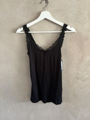 Svart spetskantad linnetopp - Snygg svart linnetopp med spetsdetaljer längs axelbanden och ringningen. Toppen har en v-ringad form och är ärmlös, vilket gör den enkel att styla till olika looks. Perfekt för dig som gillar en enkel men ändå elegant stil.