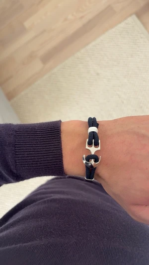 Marint armband med ankardetalj - Snyggt armband med marinblått rep och silverfärgad ankardetalj. Armbandet har en vit accent och är perfekt för dig som gillar stilrena accessoarer med nautisk känsla. Passar både till vardagslooken och när du vill addera något extra till outfiten.