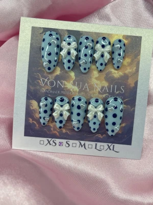 PolkaBlue (S);Medium Almond - Snygga handgjorda press-on naglar i ljusblå färg med mörkblå prickar och vita pärlmor 3D-rosetter. Naglarna har en medium almondform och är perfekta för dig som vill ha en söt och trendig look. S: 15 mm, 11,5 mm, 12 mm, 11 mm, 9 mm 