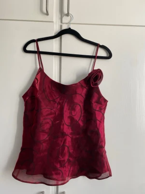 Vinröd linnetopp med rosettdetalj - Snygg vinröd linnetopp med tunna axelband och en dekorativ rosettdetalj på ena axeln, den kan tas av. Toppen har ett subtilt mönster i tyget och lätt glansig finish. 