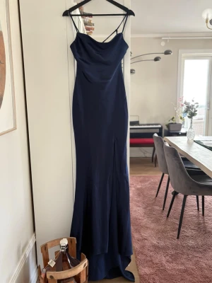 Marinblå långklänning med smala band - Elegant marinblå långklänning med smala axelband och draperad urringning. Klänningen har en figurnära passform och är tillverkad i ett glansigt, mjukt material som ger en lyxig känsla. Perfekt för speciella tillfällen när du vill känna dig extra fin. Den är använd endast en gång. Köptes för 600kr