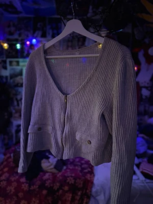 Ljusgrå ribbad kofta -  ljusgrå ribbad kofta med dragkedja framtill och två dekorativa fickor med knappdetaljer. Koftan har v-ringning och är croppad i modellen.