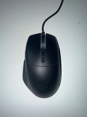 Razer Basilisk gamingmus svart - Säljer en svart Razer Basilisk gamingmus med sladd. Musen har ergonomisk form, flera programmerbara knappar och justerbart scrollhjul. Perfekt för gaming och har tydlig Razer-logga på ovansidan. Materialet är matt plast och kabeln är flätad för extra hållbarhet.