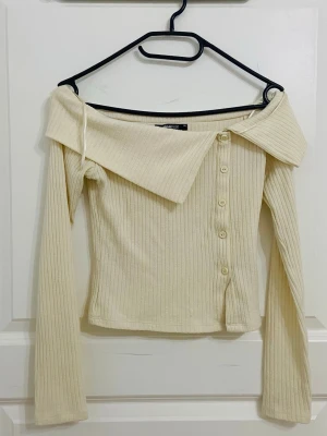 Ribbad offshoulder topp med knappar - Ribbad offshoulder topp i ljus beige/gul. Toppen har långa ärmar, dekorativa knappar framtill och en bred krage som ger en snygg omlottkänsla. Aldrig använd, säljer eftersom den inte kom till användning💕