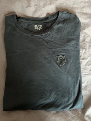 Svart EA7 t-shirt med logga - Snygg svart t-shirt från EA7 Emporio Armani med rund halsringning och en stilren logga på bröstet. Tillverkad i mjukt material som känns skönt mot huden. Perfekt för dig som gillar klassisk och enkel stil.