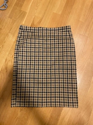 Rutig beige och svart kortkjol - Snygg kortkjol i rutigt mönster med beige, svart och bruna toner. Kjolen har en rak passform och klassisk midja. Perfekt för dig som gillar en tidlös och trendig stil. Materialet känns som bomull eller polyester.