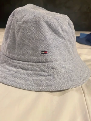 Ljusblå bucket hat från Tommy Hilfiger - Bucket hat i ljusblått från Tommy Hilfiger med diskret logotyp broderad framtill. Hatten är tillverkad i ett tunt, strukturerat bomullstyg och har en insida med blå prickar. Perfekt för att lyfta din stil under soliga dagar.