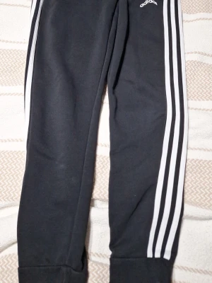 Svarta Adidas mjukisbyxor med ränder - Säljer ett par klassiska svarta mjukisbyxor från Adidas med vita signaturränder längs benen och logga vid höften. Byxorna har muddar vid bensluten och är tillverkade i mjukt, bekvämt material. Perfekta för chill eller träning.st 152