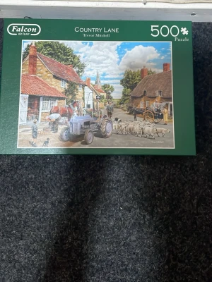 Falcon de luxe Country Lane pussel - Säljer ett 500-bitars pussel från Falcon de luxe med motivet 'Country Lane' av Trevor Mitchell. Pusslet visar en idyllisk lantlig scen med traktor, får och byhus. Perfekt för dig som gillar utmanande pussel och fina landskapsmotiv.