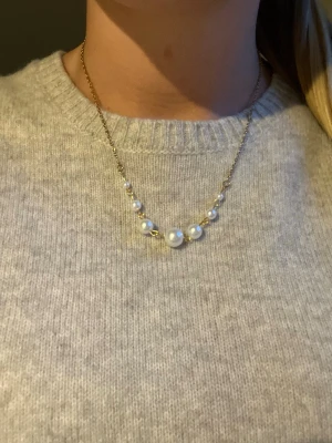 Maria Nilsdotter liknande halsband  - Snyggt halsband med tunn guldfärgad kedja och sju vita pärlor i olika storlekar. Pärlorna är placerade i mitten av halsbandet och ger en elegant och stilren look. Perfekt accessoar för att lyfta vilken outfit som helst.