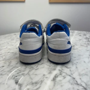 Vita och blå sneakers från Adidas - Snygga vita sneakers med blå detaljer från Adidas. Skorna har kardborreband och snörning för extra komfort och stil. De ikoniska tre ränderna i blått pryder sidorna, och Adidas-loggan syns på hälen och tungan. Relativt nya och impregnerad flera gånger. 