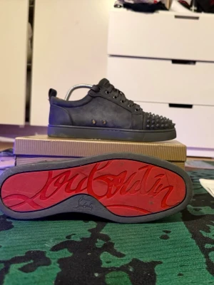 Svarta Louboutin sneakers med nitar - Säljer ett par svarta Christian Louboutin sneakers i läder med ikonisk röd sula och coola nitar på tån. Skorna har lågt skaft, svarta snören och diskreta metalldetaljer på sidan. Perfekta för dig som vill sticka ut med exklusiv streetstyle. Skorna saknar några nitar där av priset