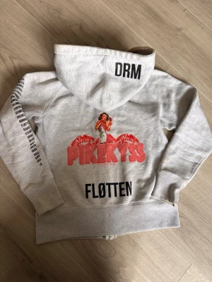 Grå hoodie med tryck från PIKEKYSS - Säljer en grå RUSS zip-hoodie från PIKEKYSS med coola tryck i rött och svart på både fram- och baksida. På ryggen finns en retroinspirerad illustration och texten 'FLØTTEN' samt 'DRM' på huvan. Ärmen har svart texttryck. Mjuk bomullsblandning och fickor fram.