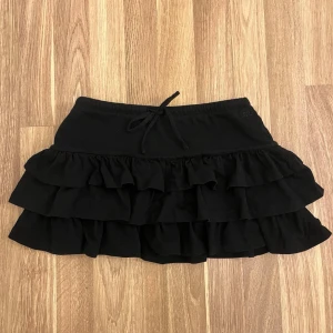 Svart volangkjol med snörning - Supersöt svart kortkjol med flera lager volanger och snörning i midjan. Perfekt för dig som gillar en lekfull och trendig stil. Kjolen har en loose passform och är tillverkad i mjukt bomullstyg.