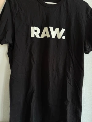Svart RAW. t-shirt från G-Star - Svart t-shirt från G-Star RAW med RAW.-tryck framtill och coola vita tryck på ryggen, inklusive club monday och NRJ. Klassisk passform med rund hals och korta ärmar. Tillverkad i mjuk bomull, perfekt för en avslappnad streetstyle.