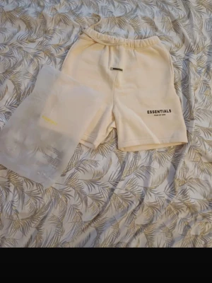 Beige shorts från Fear of God Essentials - Säljer ett par beige shorts från Fear of God Essentials med logga tryckt på framsidan. De har elastisk midja och är gjorda i mjukt bomullsmaterial. Perfekta för en avslappnad och trendig look.