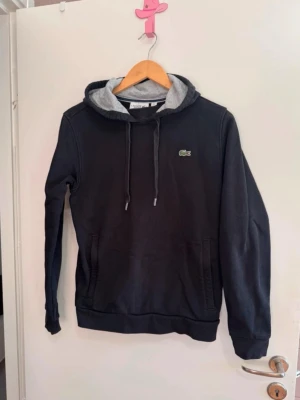 Lacoste hoodie - En svart hoodie från Lacoste sport i strl S. Grått tyg inuti luvan. Fickor i båda sidorna som stängs med dragkedja.  Tröjan är i gott skick. Den anmärkning jag kan hitta är att det släppt lite nere i ena ”flärpen” i snöret (se bild 5 + 6).