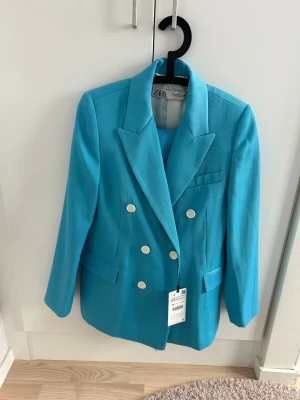 Turkost kostymset med dubbelknäppt kavaj - Snyggt turkost kostymset med dubbelknäppt kavaj och matchande byxor. Kavajen har breda slag, fyra dekorativa knappar framtill och fickdetaljer. Byxorna är raka och höga i midjan. Perfekt för dig som vill sticka ut med färg.