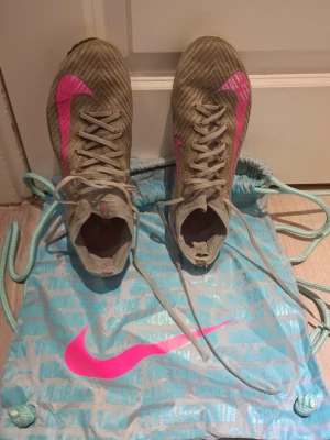 Nike Mercurial fotbollsskor beige/rosa - Säljer ett par Nike Mercurial fotbollsskor i beige med rosa detaljer och klassisk Nike-swoosh på sidan. Skorna har snörning och en strumpliknande krage för extra stöd. Ovandelen är i syntetmaterial och designen är slimmad för snabbhet på planen.