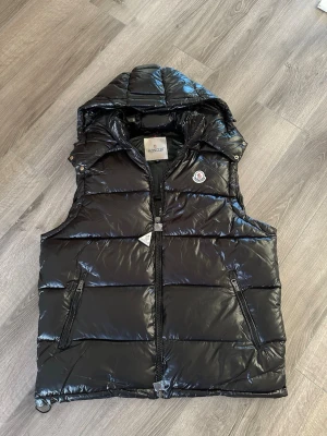 Svart moncler väst S - Snygg svart dunväst från Moncler med glansig finish och avtagbar huva. Västen har två dragkedjeförsedda fickor framtill och klassisk Moncler-logga på bröstet. Perfekt för lager-på-lager och riktigt street. Material: dun och nylon.
