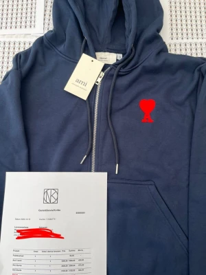 Blå zip hoodie från Ami Paris - Snygg blå hoodie med dragkedja från Ami Paris. Klassisk huva med dragsko och känguruficka framtill. På bröstet sitter Ami:s ikoniska röda hjärta med A-bokstav. Tillverkad i mjuk bomull för skön känsla. Perfekt för en avslappnad och trendig look.