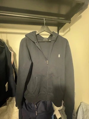 Marinblå hoodie från Polo Ralph Lauren - Snygg marinblå hoodie från Polo Ralph Lauren med dragkedja framtill och klassisk huva. Tillverkad i mjuk bomullsblandning och har fickor på magen samt den ikoniska lilla loggan broderad på bröstet. Perfekt för en chill och stilren look.