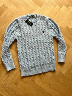 Ljusblå kabelstickad tröja Polo Ralph Lauren - Snygg ljusblå kabelstickad tröja från Polo Ralph Lauren med rund halsringning och ribbade muddar. Tröjan har den klassiska broderade loggan på bröstet och är perfekt för dig som gillar en stilren och tidlös look.