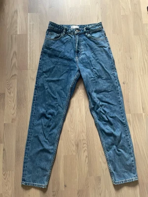 Blå raka jeans från Zara - Klassiska blå jeans från Zara med rak passform och hög midja. Jeansen har fem fickor, bälteshällor och en snygg vintage tvätt. Tillverkade i slitstark denim med en läderpatch bak i midjan. Perfekta för en avslappnad och trendig look.