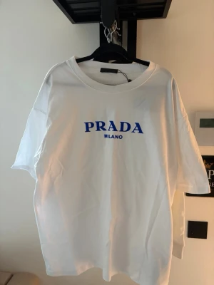 Vit t-shirt från Prada Milano - Vit t-shirt från Prada Milano med blå logotyp på bröstet. Klassisk rund halsringning och korta ärmar. Tillverkad i mjuk bomull för en skön och avslappnad look. Perfekt för dig som gillar stilrena och exklusiva plagg.