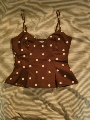 Brun prickig linnetopp med smala band - Snygg brun linnetopp med vita prickar och smala axelband. Toppen har en figurnära passform med lätt utsvängd nederdel och smock i ryggen för extra komfort. Perfekt för dig som vill ha en retroinspirerad look. Aldrig använd