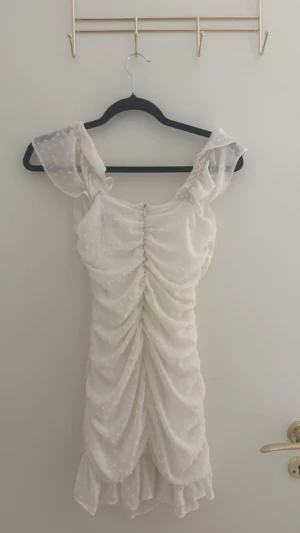 Vit kortklänning med volang och prickar - Supersöt vit kortklänning med volangdetaljer och transparenta prickiga ärmar. Klänningen har rynkade partier framtill och baktill som ger en snygg siluett. Perfekt för sommarens alla tillfällen! Axelrämmsrna har blivit omsydda