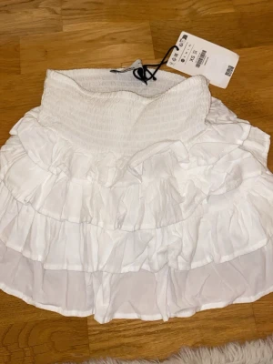 Vit volangkjol från Bershka - Supersöt vit kortkjol från Bershka med flera lager volanger och bred smock i midjan. Kjolen är gjord i ett lätt och luftigt material som passar perfekt till varma dagar. Aldrig använd, prislapp kvar.