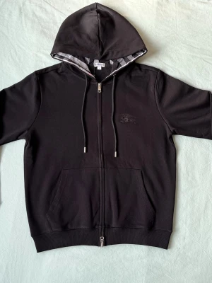 Svart zip hoodie från Burberry - Snygg svart hoodie från Burberry med dragkedja framtill och klassisk huva med rutigt foder. Hoodien har dragsko i huvan, fickor framtill och diskret Burberry-logga broderad på bröstet. Perfekt för en stilren och avslappnad look.
