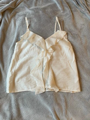 Vit linnetopp med volangdetalj - Säljer en söt vit linnetopp med tunna axelband och volang framtill. Toppen har en luftig passform och är perfekt för sommaren. Den är också nästintill oanvänd. 