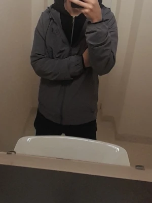 Grå vindjacka från Under Armour - Snygg grå vindjacka från Under Armour med dragkedja framtill, justerbar huva och två sidofickor. Jackan har en clean look med diskret logga på bröstet och elastiska muddar vid ärmsluten. Perfekt för blåsiga dagar och enkel att matcha med olika outfits.