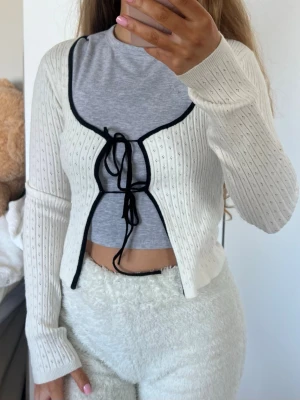 Vit ribbad kofta med svarta band - Söt vit ribbad kofta med svarta kontrastkanter och knytband framtill. Koftan har lång ärm, croppad passform. Storlek XS från FB Sister (New Yorker) endast använd nån enstaka gång 💕