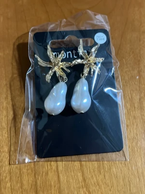 Guldörhängen med oregelbundna pärlor - Snygga örhängen från Yu&Pin med guldfärgade blommor och stora, oregelbundna vita pärlor som hänger ner. Perfekta för dig som vill sticka ut med ett unikt smycke. Materialet är metall och pärlor i en elegant design.