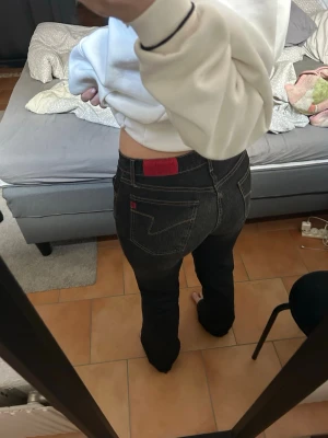 Svarta/ grå bootcut jeans - Säljer ett par svarta bootcut jeans från italien med klassiska fickor och röd logotyp på bakfickan. Jeansen har låg midja med detaljer på bak fickorna.
