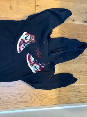 Svart Jordan hoodie med sneakersmotiv - Svart hoodie från Jordan med tryck av två färgglada sneakers och röd 'Flight'-text på bröstet. På ärmen finns det klassiska Jumpman-loggan broderad i vitt. Hoodien har huva och känguruficka framtill, perfekt för en sportig streetstyle.