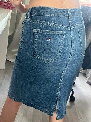 Blå jeanskjol från Tommy Hilfiger - Vintage, Snygg blå jeanskjol från Tommy Hilfiger med klassisk femficksdesign, slits bak och logga på bakfickan. Kjolen har bälteshällor och stängs med knapp och dragkedja. Perfekt för en avslappnad och trendig look. Köpt från Spanien