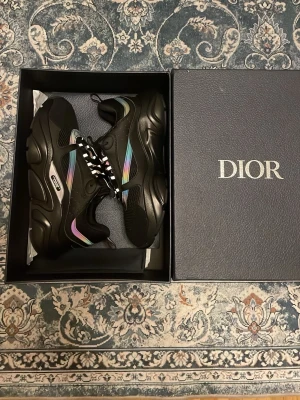 Svarta Dior sneakers med regnbågsdetaljer - Säljer ett par svarta Dior sneakers med chunky sula och regnbågsfärgade detaljer på sidan. Skorna har svartvitt mönstrade skosnören och är tillverkade i en mix av läder och textil. Perfekta för dig som vill sticka ut med en exklusiv och modern look.