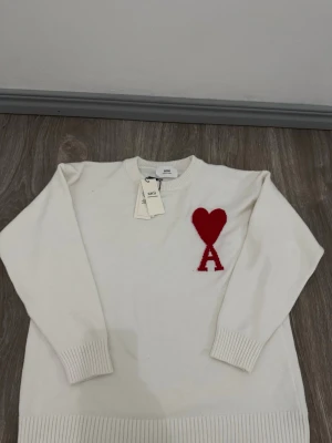 Vit stickad tröja från Ami Paris - Säljer en vit stickad tröja från Ami Paris med röd hjärtlogga och bokstaven A på bröstet. Tröjan har rund halsringning, långa ärmar och ribbade muddar vid ärmslut och nederkant. Perfekt för dig som gillar stilrena plagg med en twist.