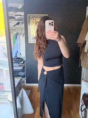 Svart långkjol från 4th & Reckless - Säljer en svart långkjol från 4th & Reckless med snygg slits framtill och en dekorativ ringdetalj i midjan. Kjolen har omlott-design och är knappt använd. 