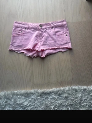 Ljusrosa jeansshorts med fransar - Säljer ett par ljusrosa jeansshorts med råa fransade kanter och klassisk femficksdesign. Shortsen har bälteshällor och knappstängning framtill. Perfekta för varma dagar och ger en avslappnad vibe.