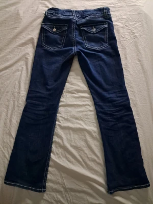 Mörkblå bootcut jeans med kontrastsömmar - Snygga mörkblå bootcut jeans med vita kontrastsömmar och dekorativa fickor bak med knappdetaljer. Klassisk femficksmodell i stretchigt denimtyg som ger en bekväm passform. Perfekta för en avslappnad och trendig look.